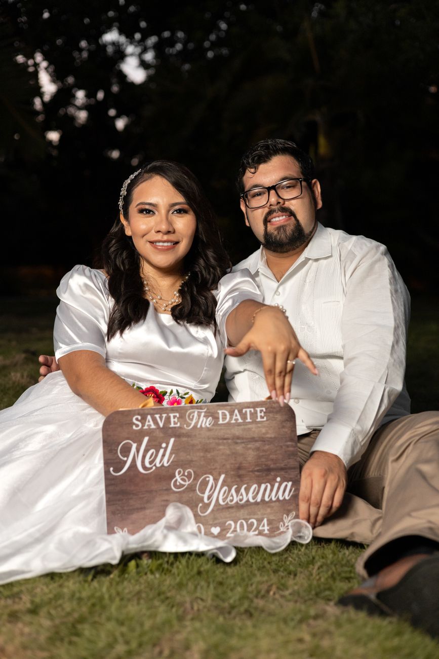 Happy couple posing with a 'Save the Date' sign in an outdoor setting during the day.